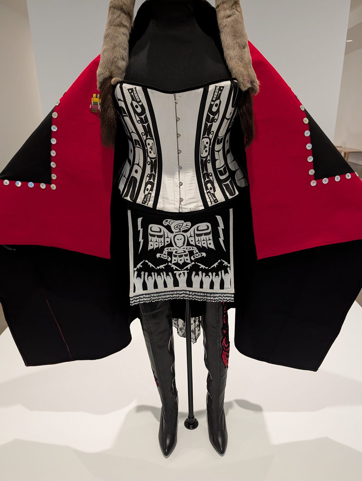 A cropped detail of Skeena Reece's "Ravens on the Colonial Fleet" depicting a bodice, cape, and high-heeled boots with first nations motifs, including the thunderbird distributing rifles to an assembled group of people. A cropped detail of Skeena Reece's "Ravens on the Colonial Fleet" depicting a bodice, cape, and high-heeled boots with first nations motifs, including the thunderbird distributing rifles to an assembled group of people.