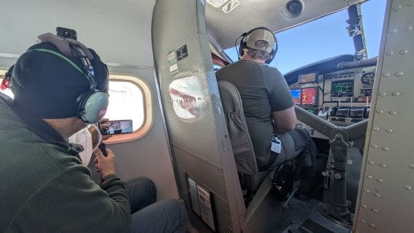 looking to the left toward a man in headphones holding a camera up to a small window and a pilot glancing out his window, his hands on his instrument panel