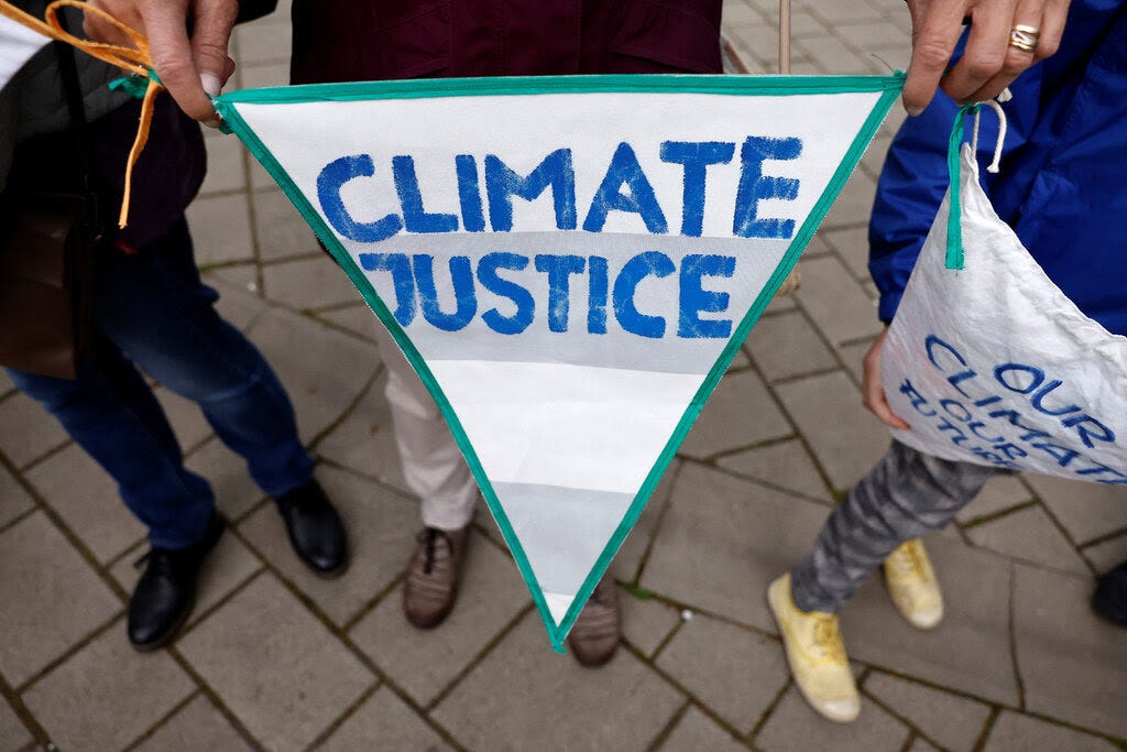 A sign reading “Climate Justice” held by three people whose legs and feet are shown against a tiled sidewalk. 