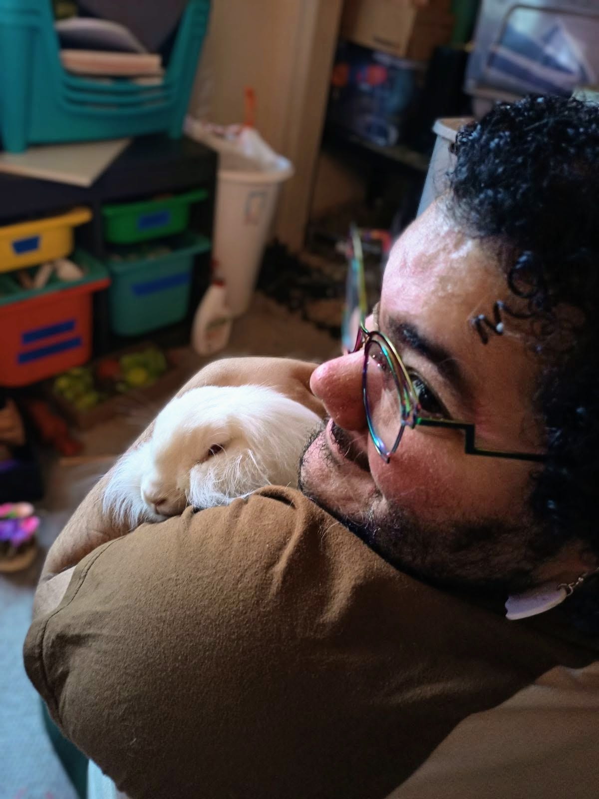 A photo of a person (me) holding a white lionhead rabbit. The camera is behind me, and I and the rabbit (Tilly) are looking at the camera over my shoulder. There is a blurry background of the room.