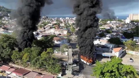 Reuters Smoke rises from burning busses in Mexico. We can see a scene of the city from a distance with sea and blue skies in teh bcakground