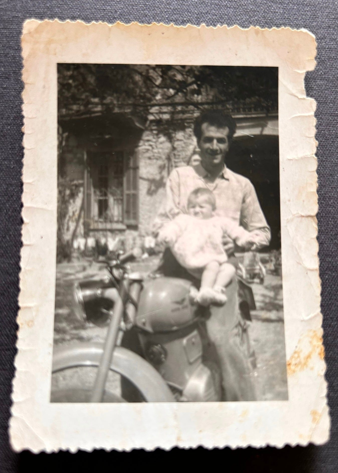 Black and white photograph from approximately 1957 showing a young father seated on his Moto Guzzi motorcycle with an infant on his lap, taken in northern Italy. A moment of quiet pride and joy in modest but dignified surroundings.