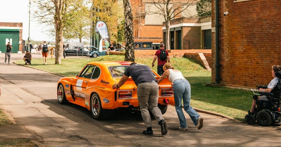 People pushing a bright orange vintage race car
