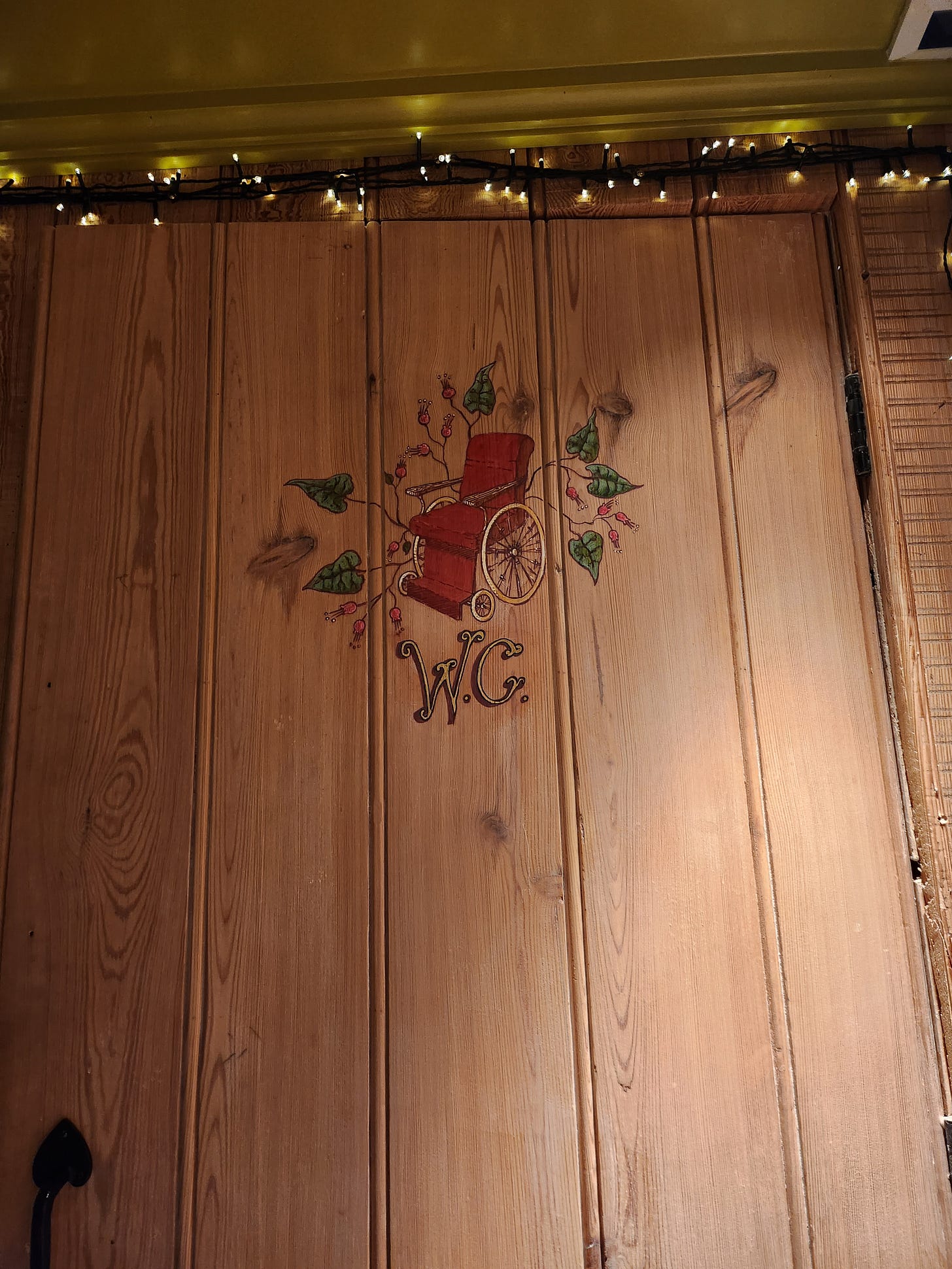 On a wooden door, an antique wheelchair is painted, surrounded by ivy, to indicate that this bathroom is wheelchair friendly