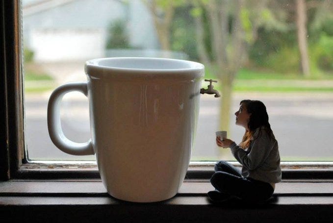 A miniature woman with brown hair wearing a gray sweater and jeans sits cross-legged on a wooden windowsill holding a small white cup near her face while looking at a enormous white ceramic coffee mug positioned outside the window with a faucet attached to its side pouring a stream of liquid into the mug the background includes green grass a tree and a street the scene emphasizes scale contrast between the tiny figure and the oversized object