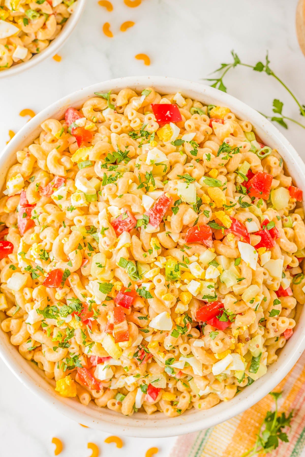 A bowl of macaroni salad with diced tomatoes, yellow bell peppers, chopped eggs, and herbs on a white surface. A bowl of macaroni salad with diced tomatoes, yellow bell peppers, chopped eggs, and herbs on a white surface.