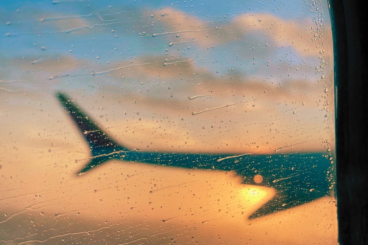 The wing of an airplane seen through a rainy window at sunset, creating a blurry, moody image with warm, orange light. The wing of an airplane seen through a rainy window at sunset, creating a blurry, moody image with warm, orange light.