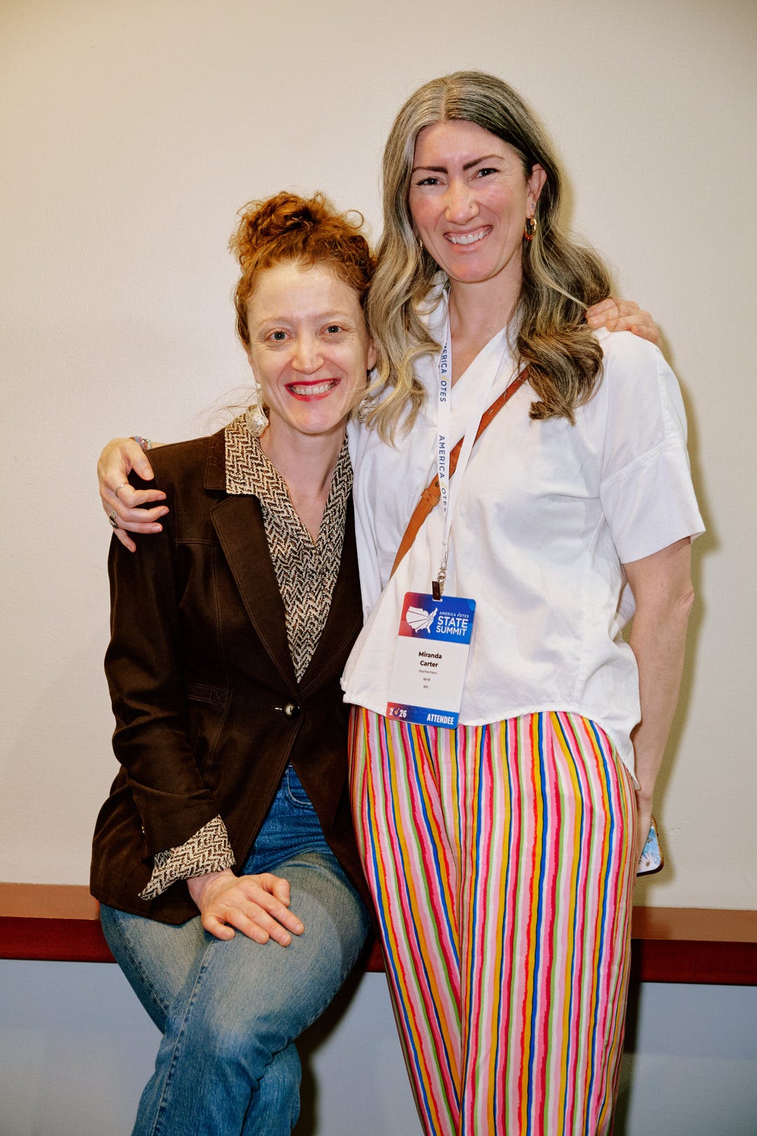 Two women leaning against a wall with arms around each other, one in a brown blazer with copper red hair, one in a white top and rainbow striped pants with a Miranda Carter badge