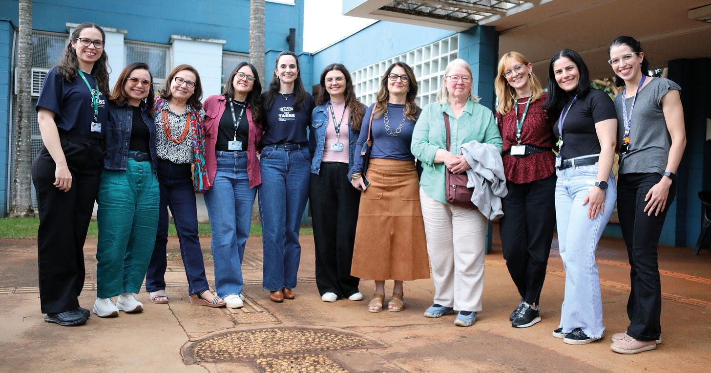 Onze pessoas em pé enfileiradas em um pátio de instituição com paredes azuis, piso de concreto com padrão geométrico, palmeira ao fundo, algumas vestindo crachás de identificação, com roupas em cores variadas incluindo tons de azul, preto, rosa, verde e marrom, a maioria usando óculos, em ambiente externo com cobertura parcial e estrutura arquitetônica ao fundo. Onze pessoas em pé enfileiradas em um pátio de instituição com paredes azuis, piso de concreto com padrão geométrico, palmeira ao fundo, algumas vestindo crachás de identificação, com roupas em cores variadas incluindo tons de azul, preto, rosa, verde e marrom, a maioria usando óculos, em ambiente externo com cobertura parcial e estrutura arquitetônica ao fundo.