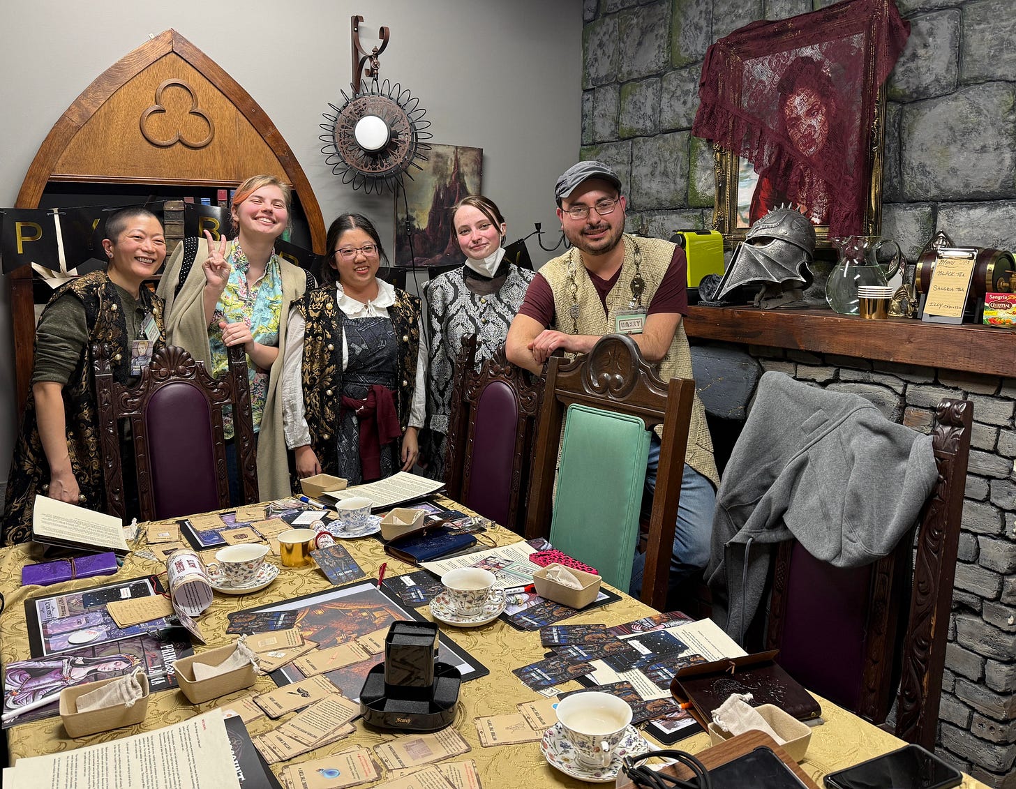 Foreground: table with cards and gaming accessories. L to R: Logan, Blonde female smiling and making the peace sign, asian female with glasses, white woman in a black lacey top smiling, and a white man with a gray hat and glasses. If you know, you will know that zora is represented in the portrait on the wall, veiled by a lacey maroon shawl.