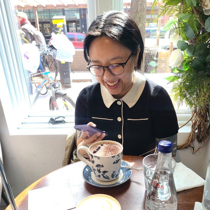 Two images side by side, on the left is K wearing a cute black and white short-sleeved shirt, sitting at a table in a coffee shop in front of a window, giving the camera the middle finger with a cheeky expression. On the right is the same scene, but K is smiling down at their phone.