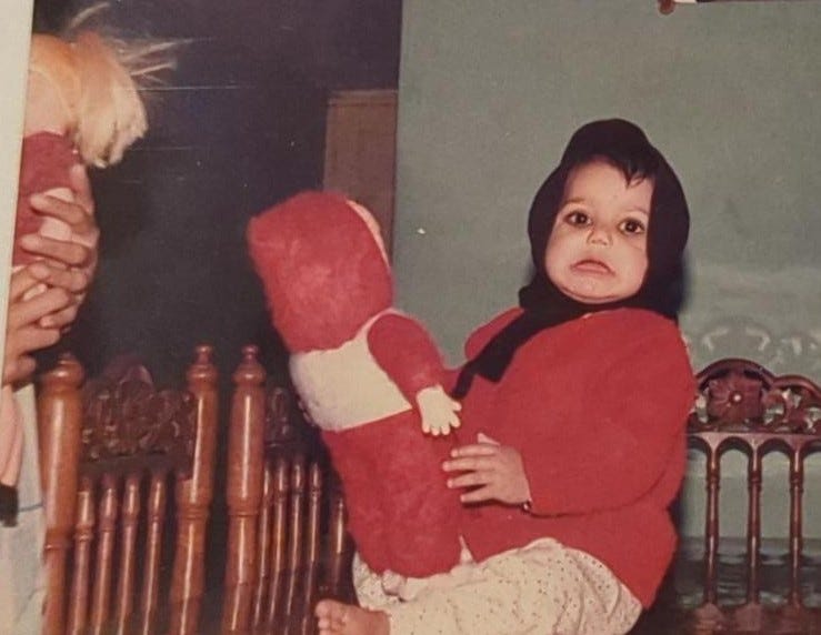 1-year-old baby in red sweater and black scarf on her head, seated on a wooden dining room table, with a dismayed expression. She's holding a white doll in a red outfit.