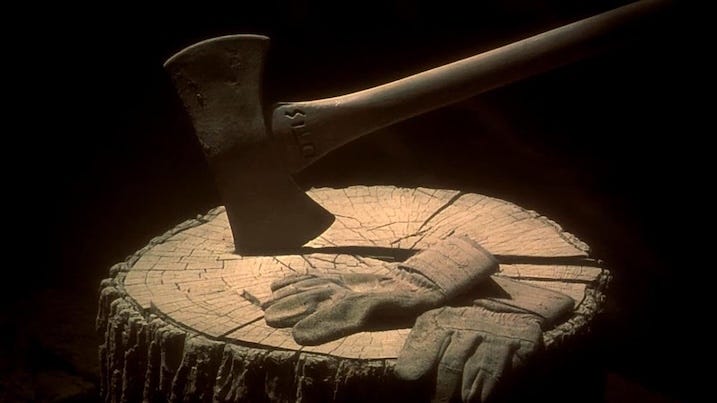 The axe "Otis" embedded in a wooden stump, resting beside a pair of worn work gloves under dramatic lighting.