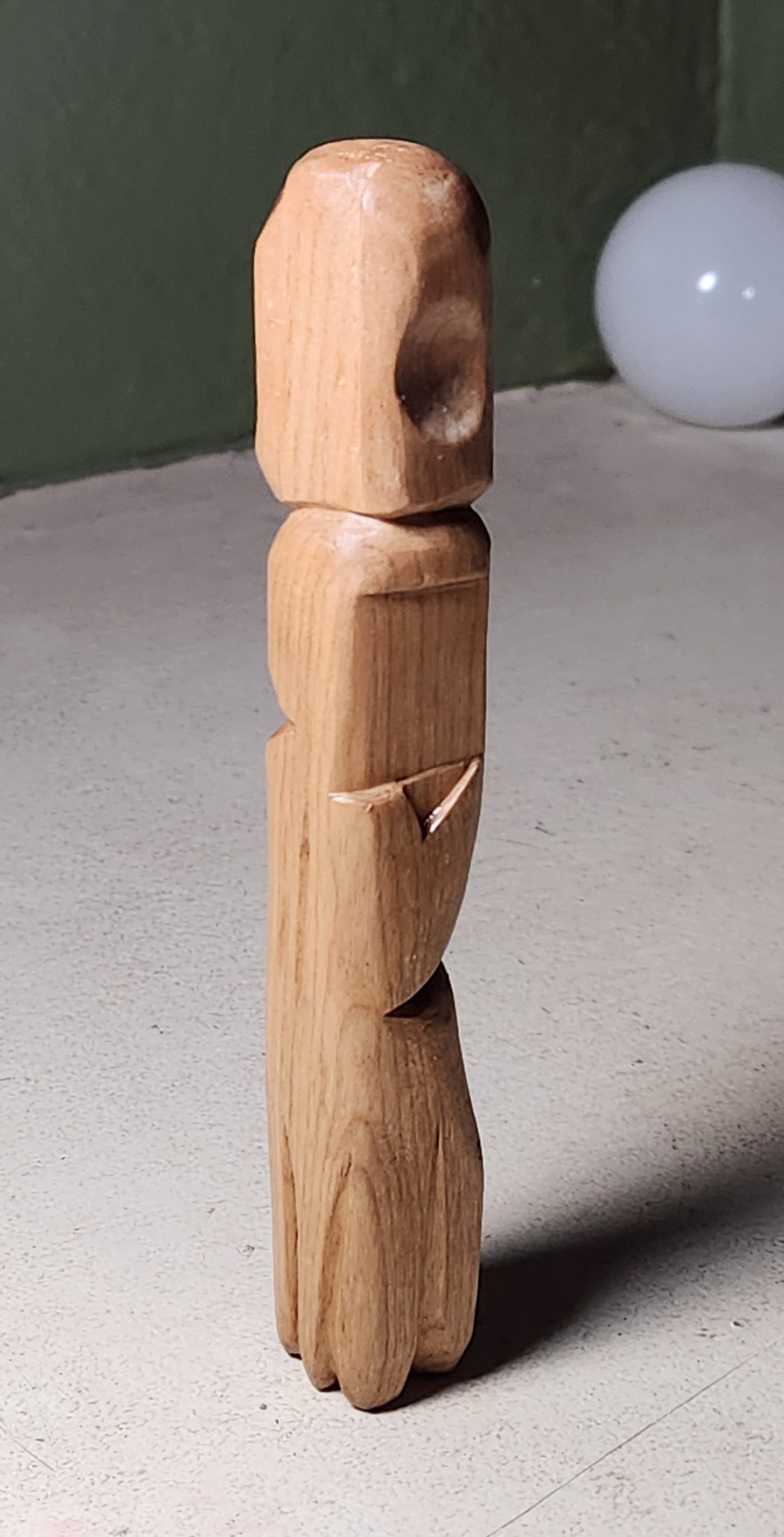 A photograph of a woodcarving - tall and thin, approximately square in cross-section, carved semi-abstractly. The carving is on a gray-painted concrete surface in front of an olive-painted cinderblock wall, with a glass globe in the distance.