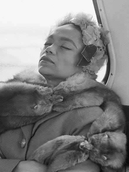 This may contain: a woman wearing a fur coat and flower in her hair is sitting on a bus