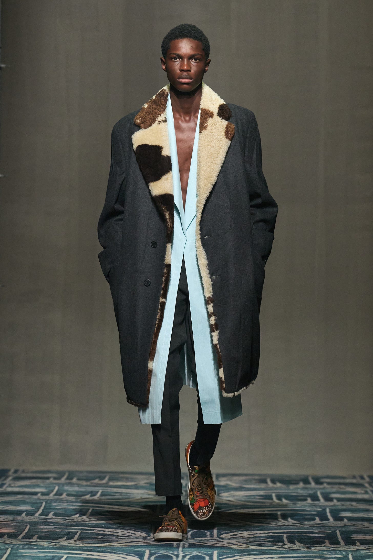 A model walks the runway wearung a charcal gray overcoat with a shearling collar over an ice blue shirt.