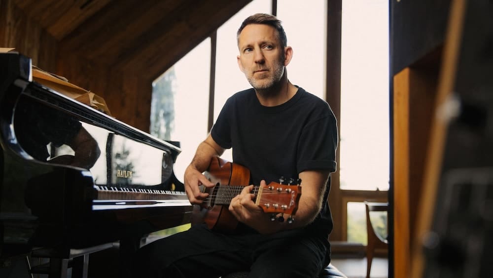 Denison Witmer holding an acoustic guitar while sitting at a piano