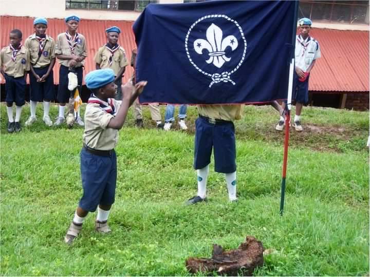 A group of young men standing next to each other in front of a building. An image of young boy scouts holding up a flag on top of a grass field. A group of young scouts walking down a dirt road. A group of young people walking down a street holding a sign with the words 'scout peace'.