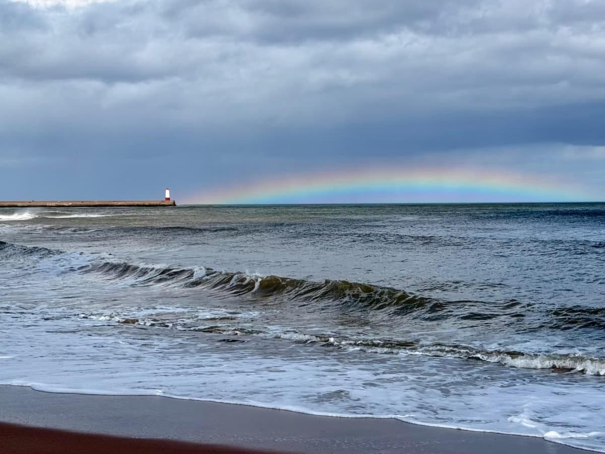 A photo taken from a beach looking at a rainbow coming from the Berwick-upon-Tweed lighthouse.
