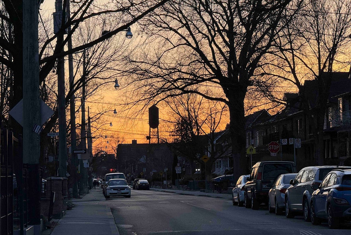 A street scene at dusk with a clear sky transitioning from orange to blue. The street is lined with parked cars on both sides and a few cars driving down the road. Bare trees with no leaves are visible, and streetlights hang from wires above the street. There are houses and buildings along the sides of the street, and a stop sign is visible on the right side of the image. The image captures the calm and quiet atmosphere of a residential neighborhood during sunset.