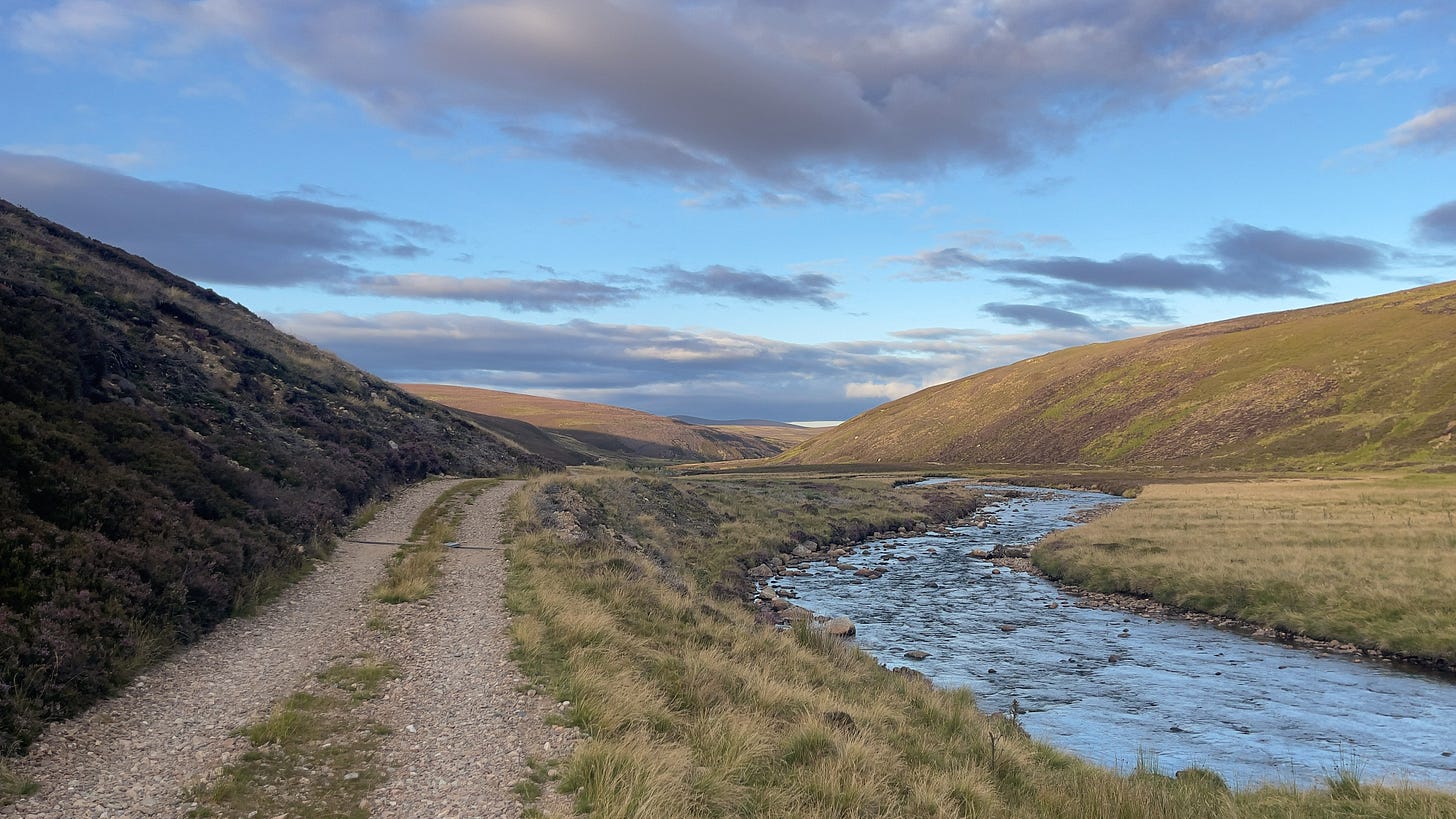 A gentle trail descends alongside winding rivers that carve through rolling hills. The sky is wide and bright, and the river’s surface glows with golden reflections from the evening sun, creating a serene, almost luminous landscape.
