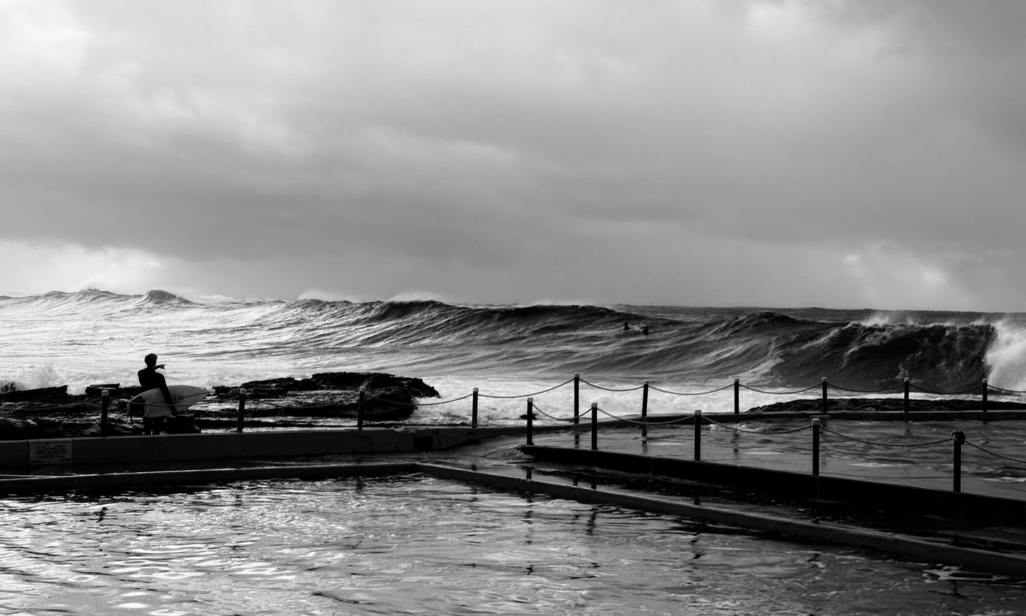 Black and white image of cresting wave breaking into a barrel roll with a surfer in the foreground standing on the rocks watching it break by an ocean pool.