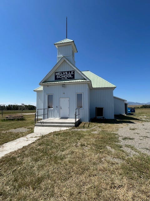 White wooden building resembling a church is a country schoolhouse in Melville, Montana. The Sweetgrass River is shallow, rocky, calm, and shady.