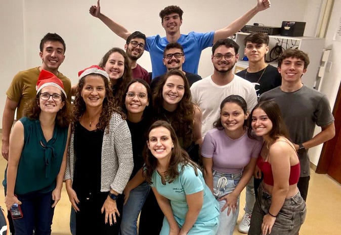 Uma fotografia mostra um grupo de cerca de 15 pessoas sorrindo em uma sala fechada. Algumas usam gorros vermelhos de Natal, enquanto outras estão com roupas casuais. Um homem aparece parcialmente no canto esquerdo, tirando a selfie. Ao fundo, um rapaz ergue os braços em sinal de comemoração, criando um clima de descontração e confraternização Uma fotografia mostra um grupo de cerca de 15 pessoas sorrindo em uma sala fechada. Algumas usam gorros vermelhos de Natal, enquanto outras estão com roupas casuais. Um homem aparece parcialmente no canto esquerdo, tirando a selfie. Ao fundo, um rapaz ergue os braços em sinal de comemoração, criando um clima de descontração e confraternização