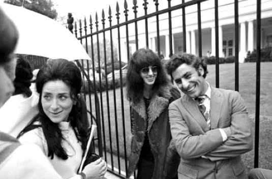 A woman who clearly looks like a hippy/rock star type leaning on the fence on the White House smiling. She’s wearing what looks like a suede coat with fur lining/collars. Next to her is a smiling man in a suit, who would look more respectable than hair were it not for his palpable air of mischief