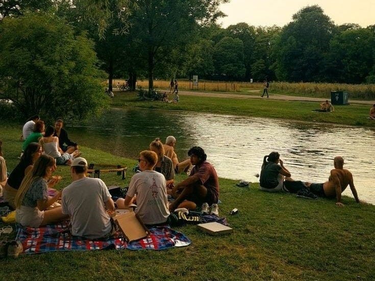 This may contain: many people are sitting on the grass next to a river and picnicking at sunset