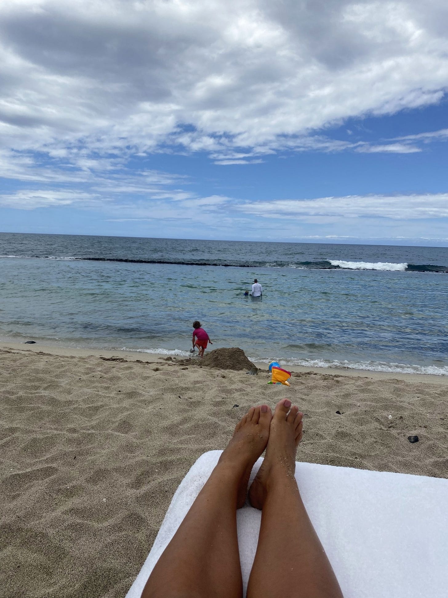The view from a beach lounger in Hawaii, bare feet propped up, ocean and a child playing at the shoreline.