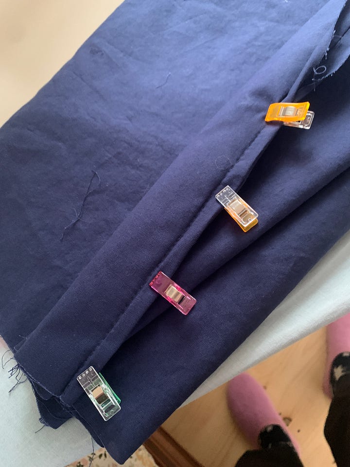 first photo is a knitted rectangle of multicolored stripes, still on the knitting needles, sitting on a wooden table. the second photo is a straight seam sewn in navy blue cloth, with four quilting clips holding it in place