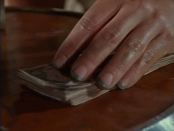 A close-up of a white hand touching a stack of cash. The hand contains one of the dirtiest sets of fingernails in recorded history.
