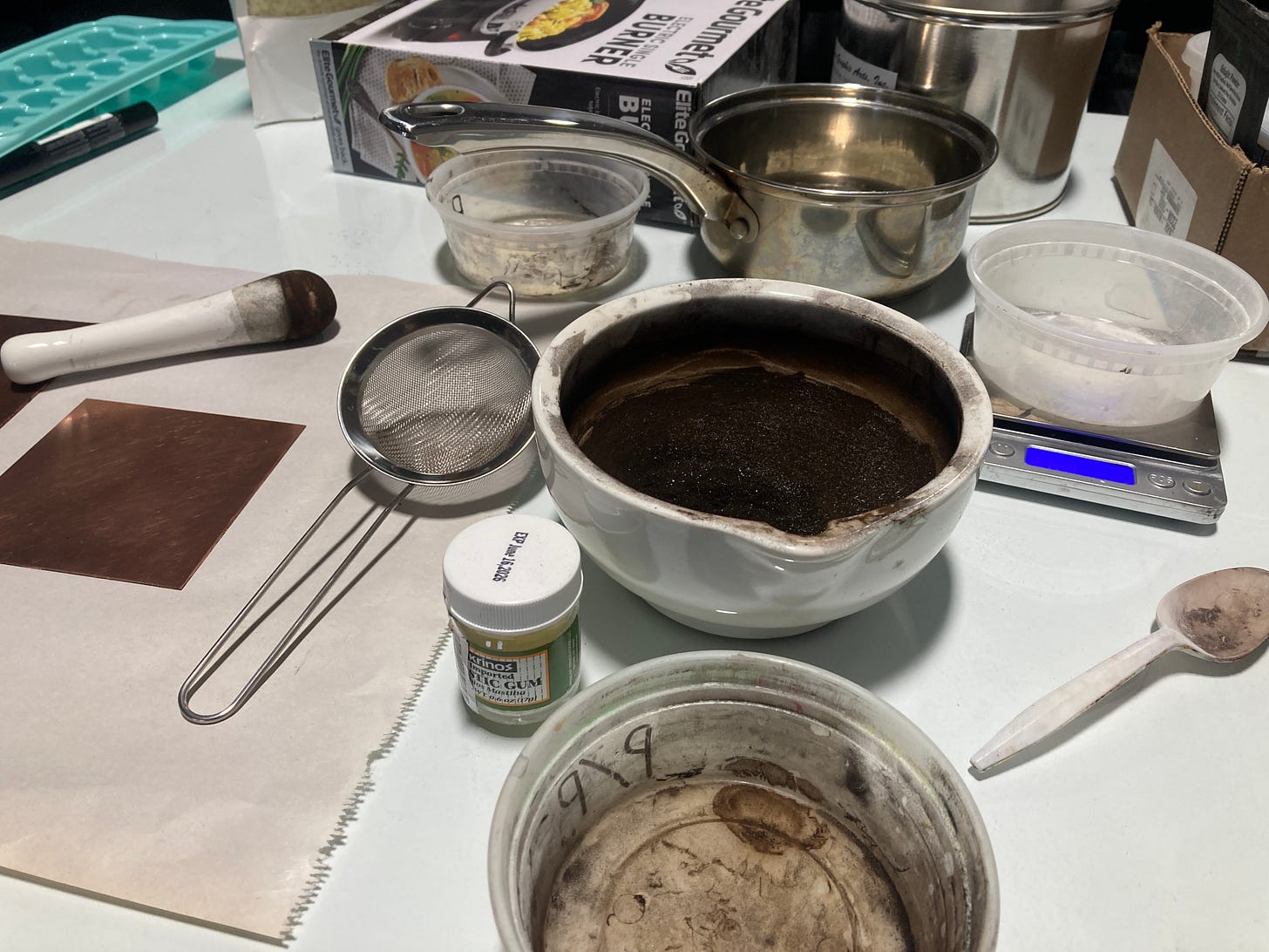An assortment of containers, including a mortar and small cooking pot, holding various ingredients needed to make hard ground rest on a white surface. There is also a small copper plate, metal sieve, plastic spoon, and ice cube tray.