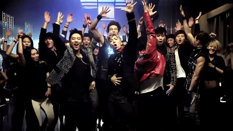 A group of K-pop idols mostly in dark clothes jump around with their hands in the air. A group of K-pop idols mostly in dark clothes jump around with their hands in the air.