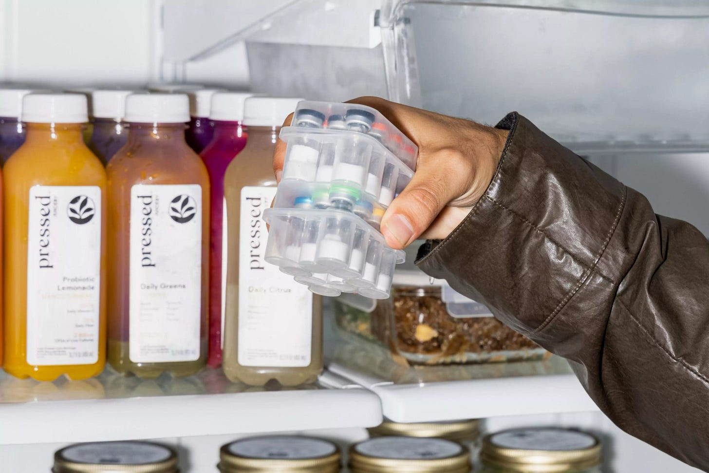 A hand in a brown leather jacket holds two stacked pill organizers inside a fridge next to bottles of pressed juice labeled Daily Greens and Probiotic Lemonade.