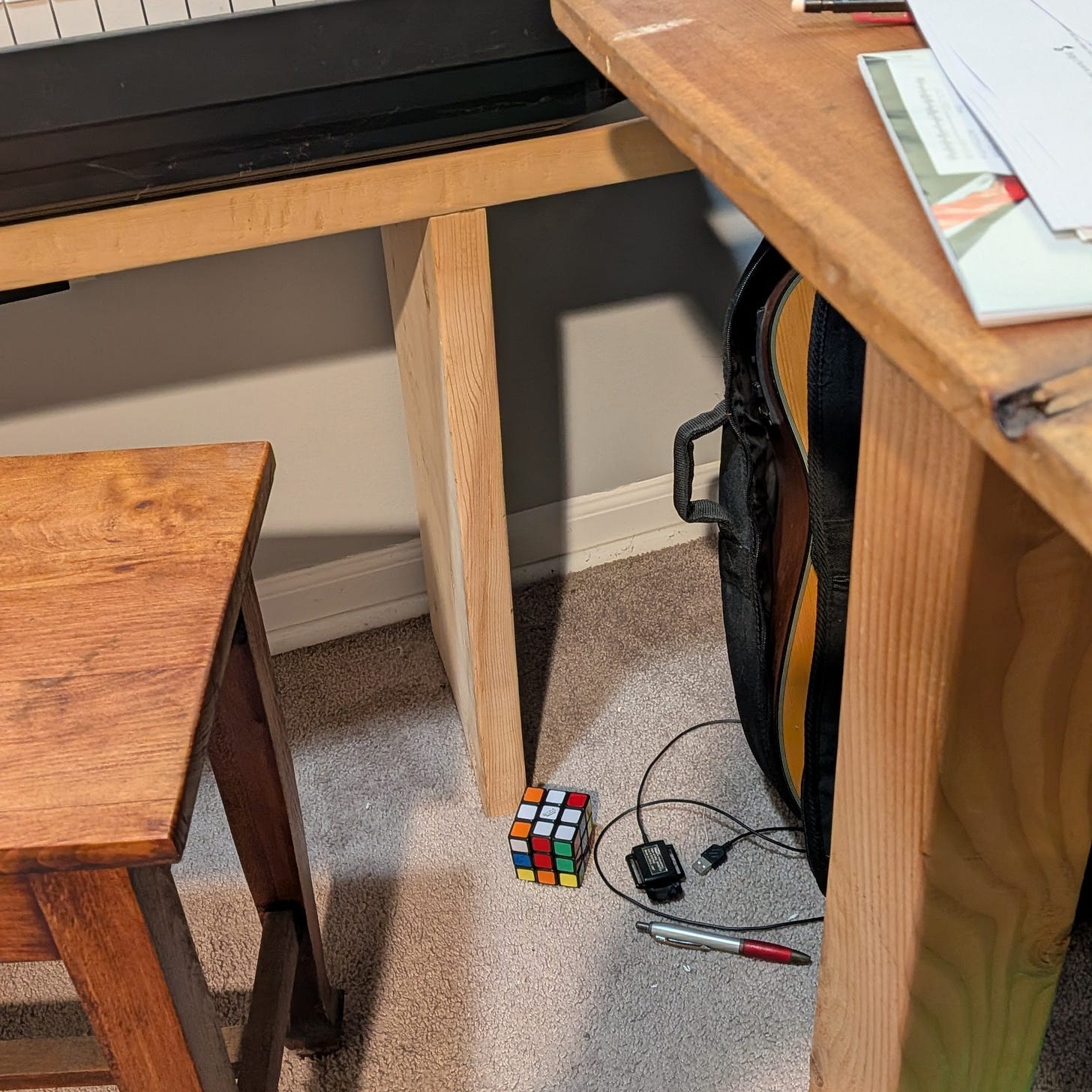 A medium shot shows a scrambled Rubik's Cube on a gray carpet floor beneath a wooden desk. Next to it are a black USB cord with a connector and a pen with a dark red grip and silver top. In the background on the right is a black gig bag containing a stringed instrument. A medium shot shows a scrambled Rubik's Cube on a gray carpet floor beneath a wooden desk. Next to it are a black USB cord with a connector and a pen with a dark red grip and silver top. In the background on the right is a black gig bag containing a stringed instrument.