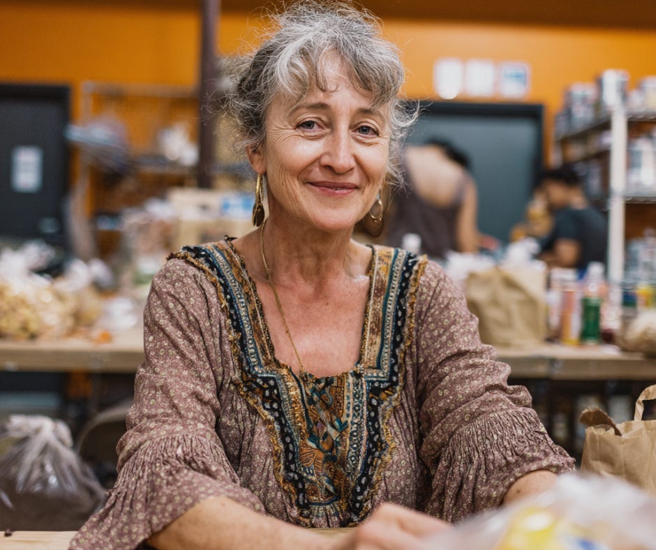 A smiling older woman with gray hair and boho style clothing sitting at a table during a creative workshop, surrounded by art supplies and materials.
