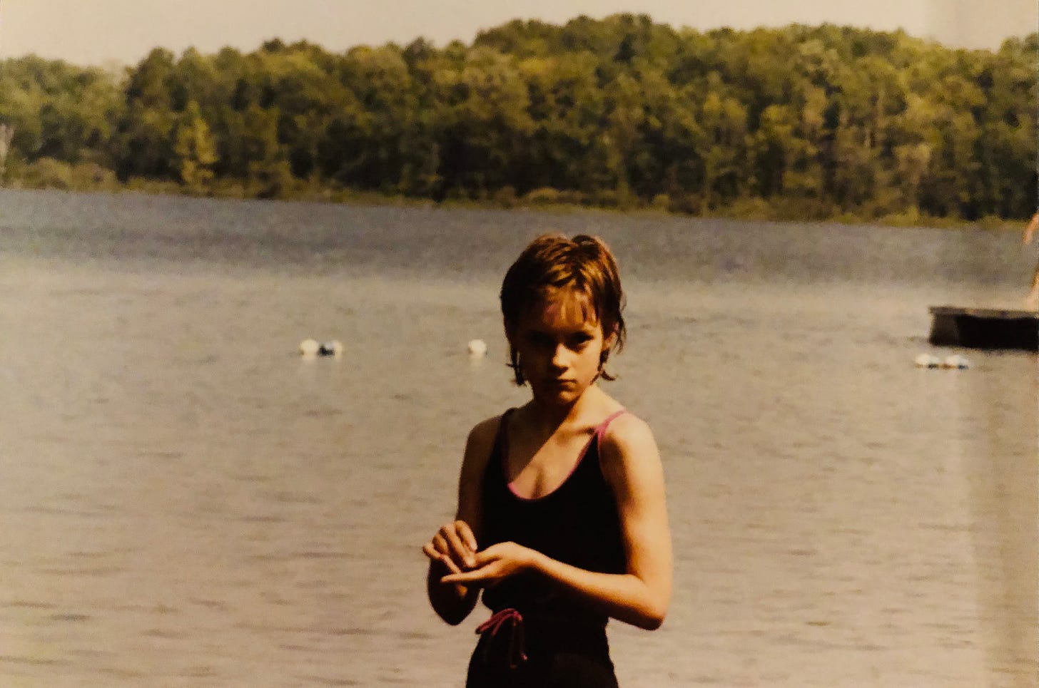 A grouchy twelve-year-old girl in a black swimsuit glares at you from the shore of a lake with swimming buoys and trees in the background A grouchy twelve-year-old girl in a black swimsuit glares at you from the shore of a lake with swimming buoys and trees in the background