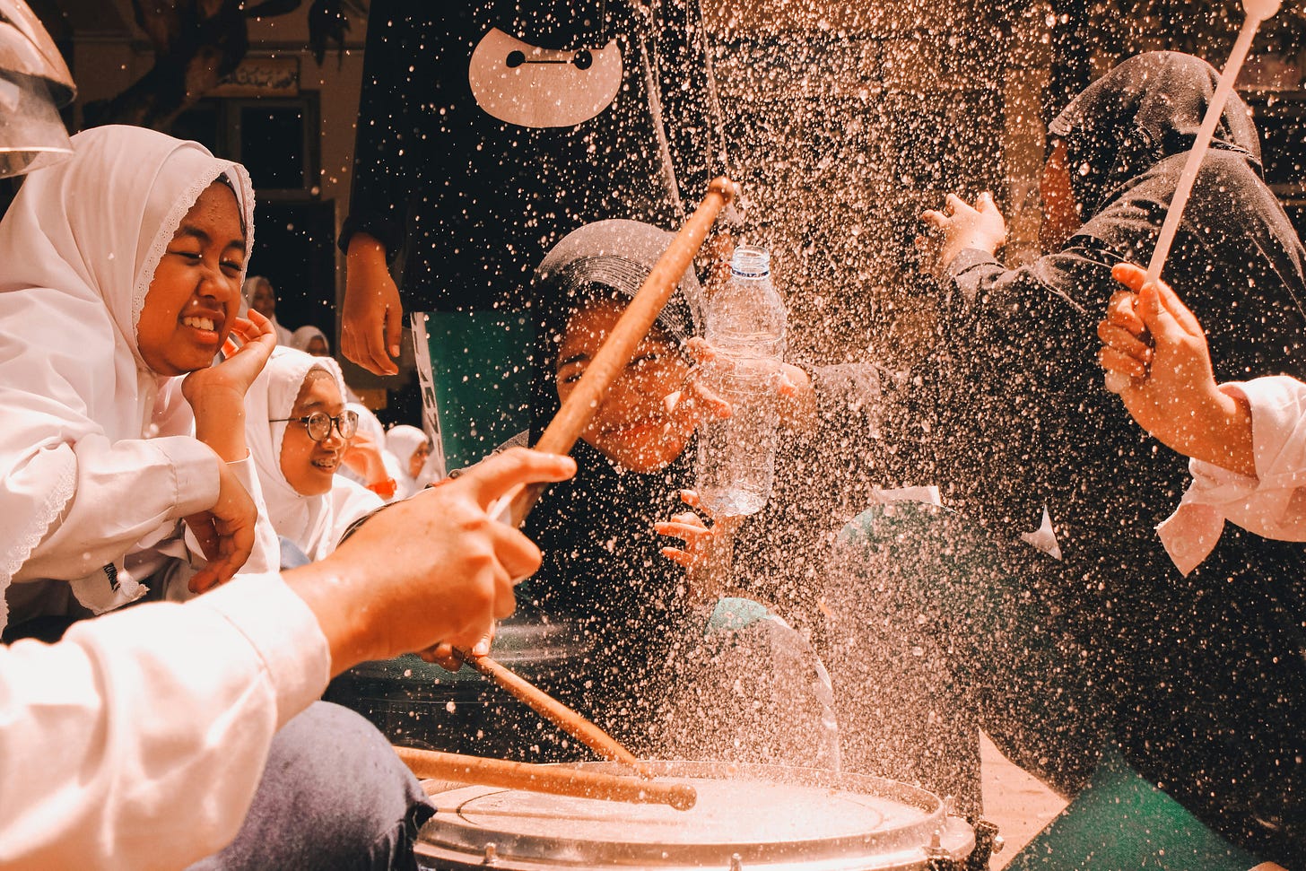 A candid, warm-toned action photograph captures Indonesian schoolgirls wearing white hijabs and uniforms, laughing and cheering as water splashes dramatically over a snare drum. Hands in the foreground enthusiastically beat the drum sticks into the splashing water, evoking a sense of vibrant youth, joy, and shared local culture. The scene encapsulates the lively spirit of the Indonesian people, reflecting a nation celebrated for its rich cultural diversity and more than 700 local languages. A candid, warm-toned action photograph captures Indonesian schoolgirls wearing white hijabs and uniforms, laughing and cheering as water splashes dramatically over a snare drum. Hands in the foreground enthusiastically beat the drum sticks into the splashing water, evoking a sense of vibrant youth, joy, and shared local culture. The scene encapsulates the lively spirit of the Indonesian people, reflecting a nation celebrated for its rich cultural diversity and more than 700 local languages.