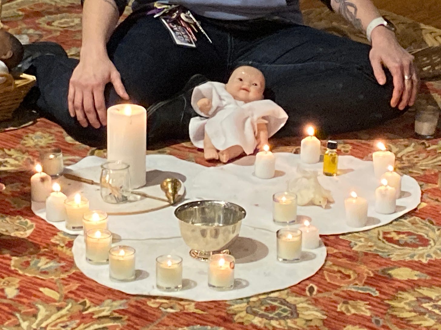 The Godly Play baptism story set out with the bowl of water, Christ candle, dove, and chrism, surrounded by lit candles. A baby doll is propped against a person in the background.