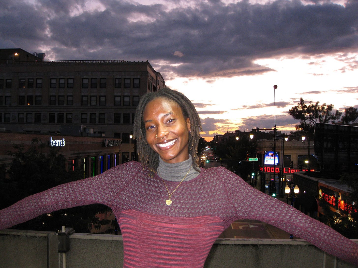 A photo of me with the city in the background. I am wearing a gray turtle neck with a burgandy mesh top over it. I am smiling with my arms out to my sides. It is cloudy in the back and the sunset is in the horizon line.