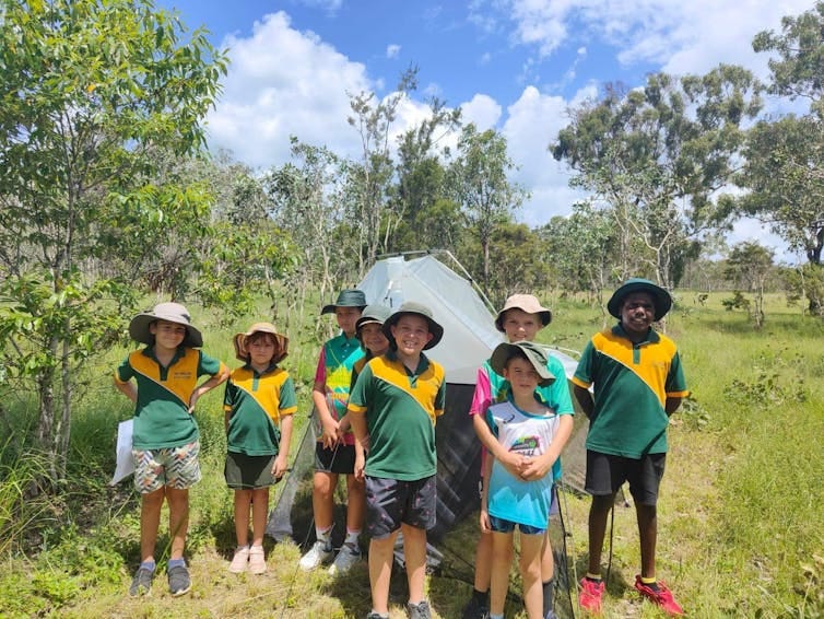 Seven children in green and yellow school uniforms stand outside in front of an insect trap. Seven children in green and yellow school uniforms stand outside in front of an insect trap.