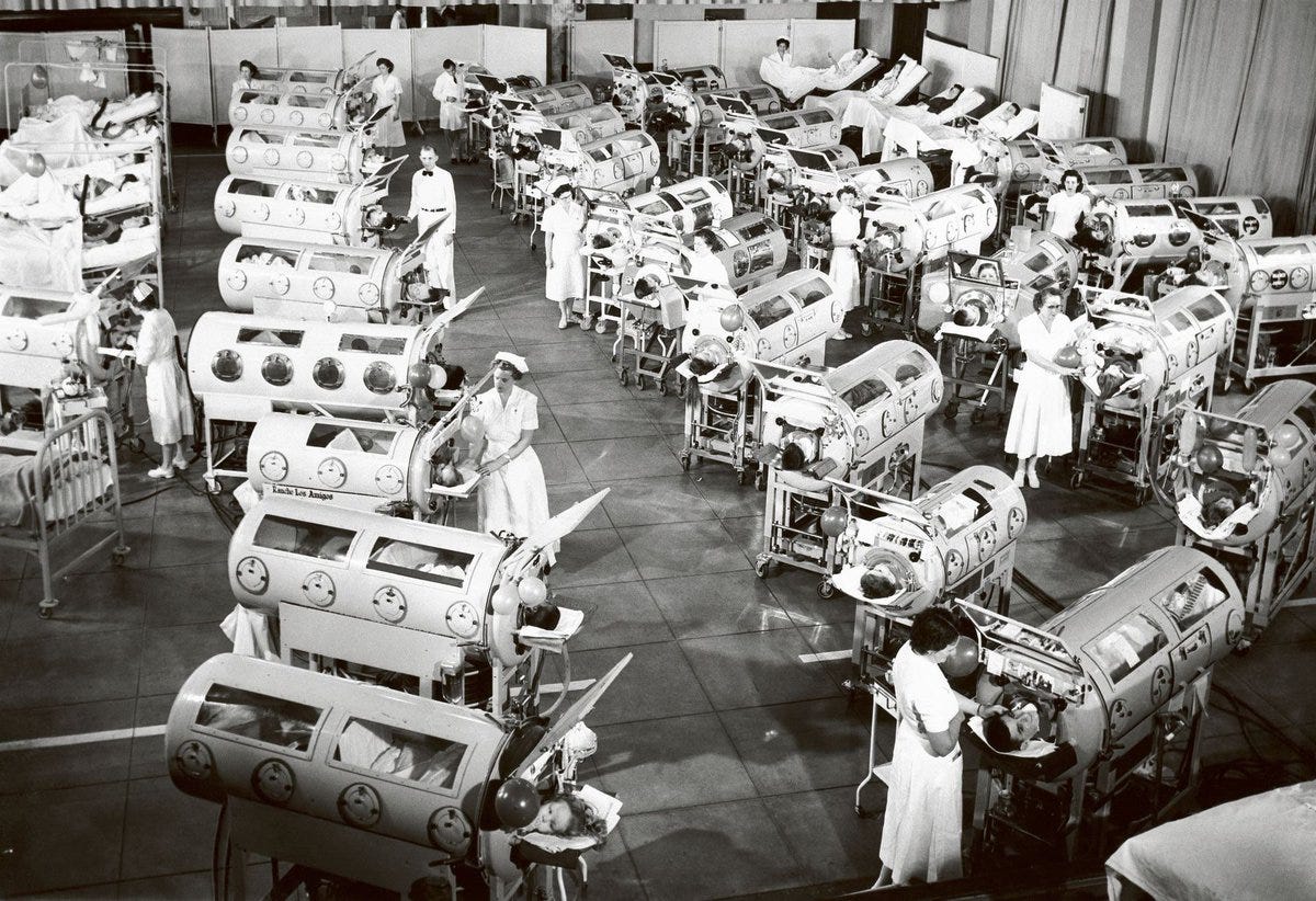 A black-and-white photo of a large room filled with numerous iron lungs, cylindrical medical devices used for polio treatment. Medical staff in white uniforms stand and work around the machines, attending to patients inside. The room is spacious with a tiled floor and multiple rows of iron lungs, each with visible portholes and mechanical components. A black-and-white photo of a large room filled with numerous iron lungs, cylindrical medical devices used for polio treatment. Medical staff in white uniforms stand and work around the machines, attending to patients inside. The room is spacious with a tiled floor and multiple rows of iron lungs, each with visible portholes and mechanical components.