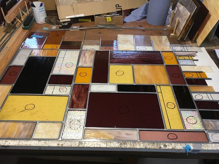 Stained glass panel work in progress, made up of multiple rectangular and square panels of different colours and textures. The colours include brown, yellow, orange, maroon and clear glass.