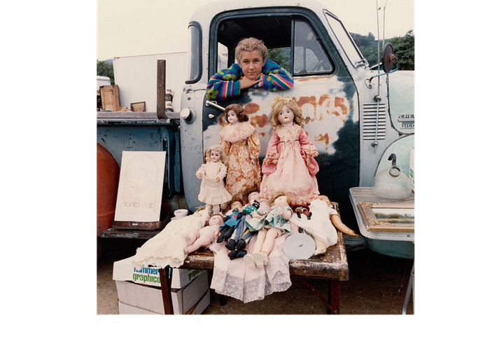 A girl in a vintage truck with vintage dolls, two men sitting on a car, one man with dreadlocks in front of a hatchback and a guitar