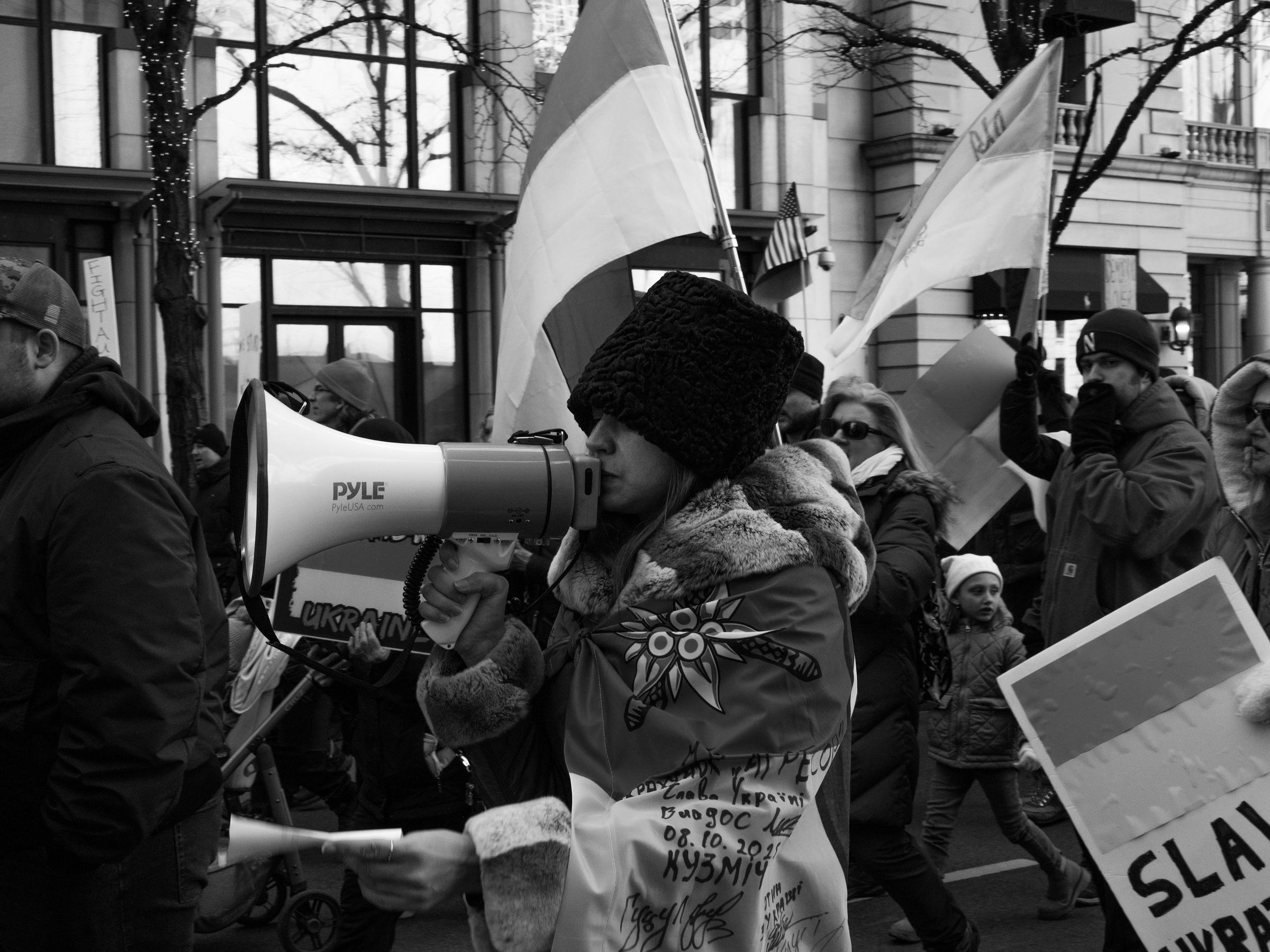 A black and white photograph captures a protester passionately speaking into a megaphone while marching. They wear a fur-lined coat with embroidered details and a thick, furry hat. Behind them, Ukrainian flags wave as a diverse crowd, including children, marches in support. Signs with bold lettering and determined expressions emphasize the urgency of their message. Reflections in glass buildings add depth to the urban setting. A black and white photograph captures a protester passionately speaking into a megaphone while marching. They wear a fur-lined coat with embroidered details and a thick, furry hat. Behind them, Ukrainian flags wave as a diverse crowd, including children, marches in support. Signs with bold lettering and determined expressions emphasize the urgency of their message. Reflections in glass buildings add depth to the urban setting.