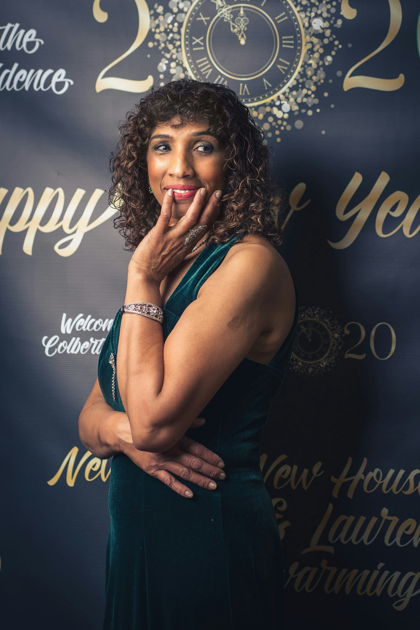 Confident Black woman in emerald green velvet dress posing elegantly at New Year's event, wearing curly hair and a sparkling bracelet, with a decorative gold clock and '2024' backdrop