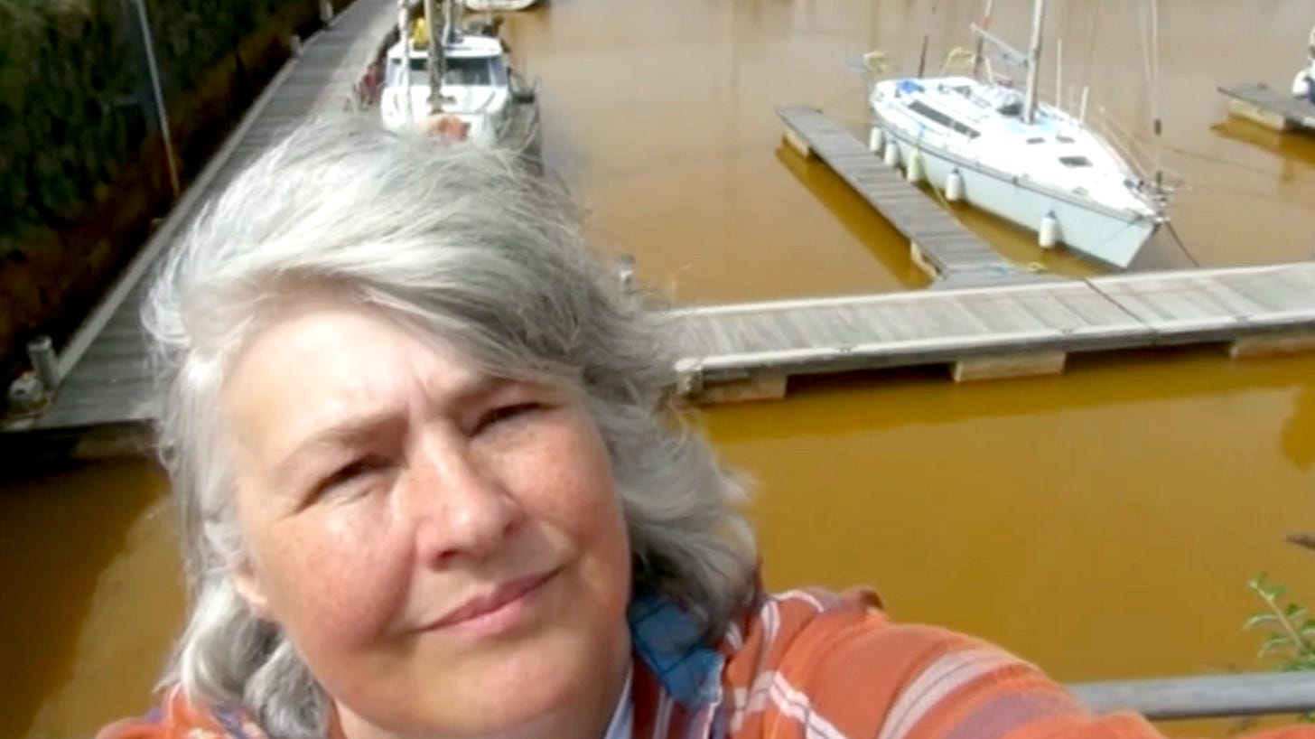 Marianne Birkby is taking a selfie in front of the orange water at Whitehaven Harbour. The harbour has a wooden pontoon, and two boats docked. Birkby has mid-length grey hair and is wearing an orange jacket. Marianne Birkby is taking a selfie in front of the orange water at Whitehaven Harbour. The harbour has a wooden pontoon, and two boats docked. Birkby has mid-length grey hair and is wearing an orange jacket.