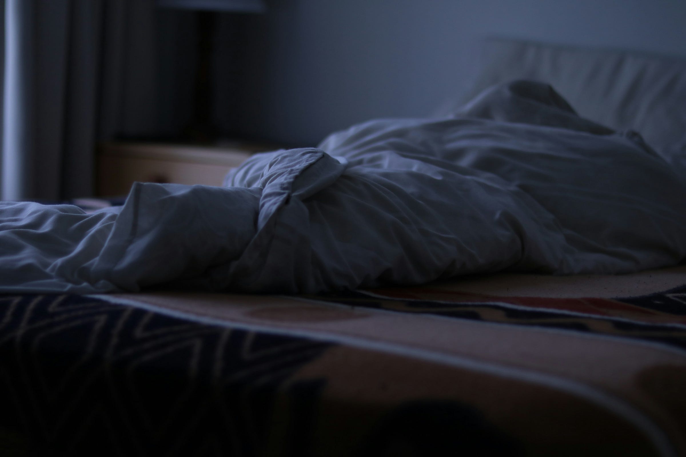 The photo of a dimly lit bedroom showing an unmade bed with a white blanket twisted in the center, suggesting recent use. A pillow rests near the headboard, and soft light filters through a window in the background.